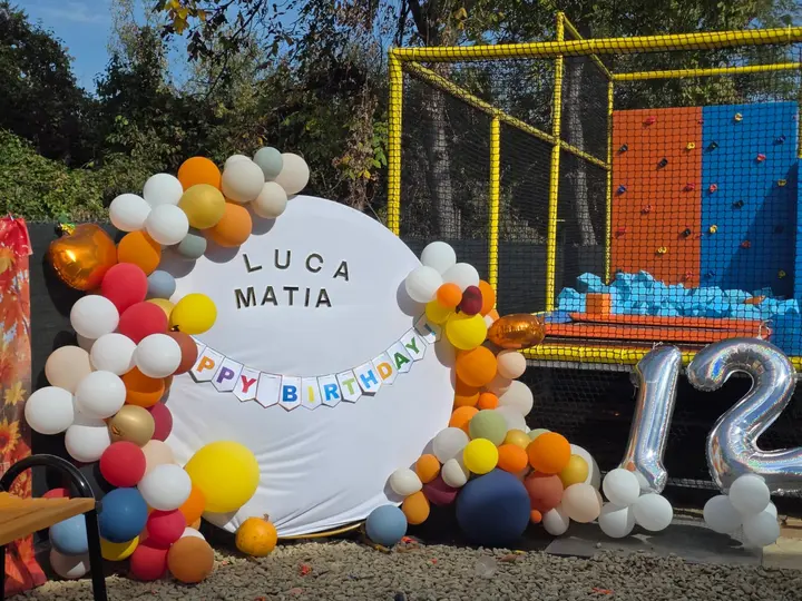 Panou aniversar cu numele copiilor, arcadă de baloane colorate și cifra 12, lângă arena de trambuline